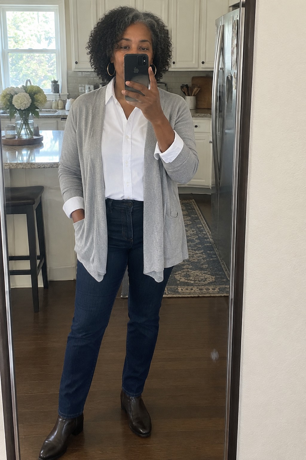 Woman with curly gray hair in white shirt, gray cardigan, and jeans in kitchen mirror selfie