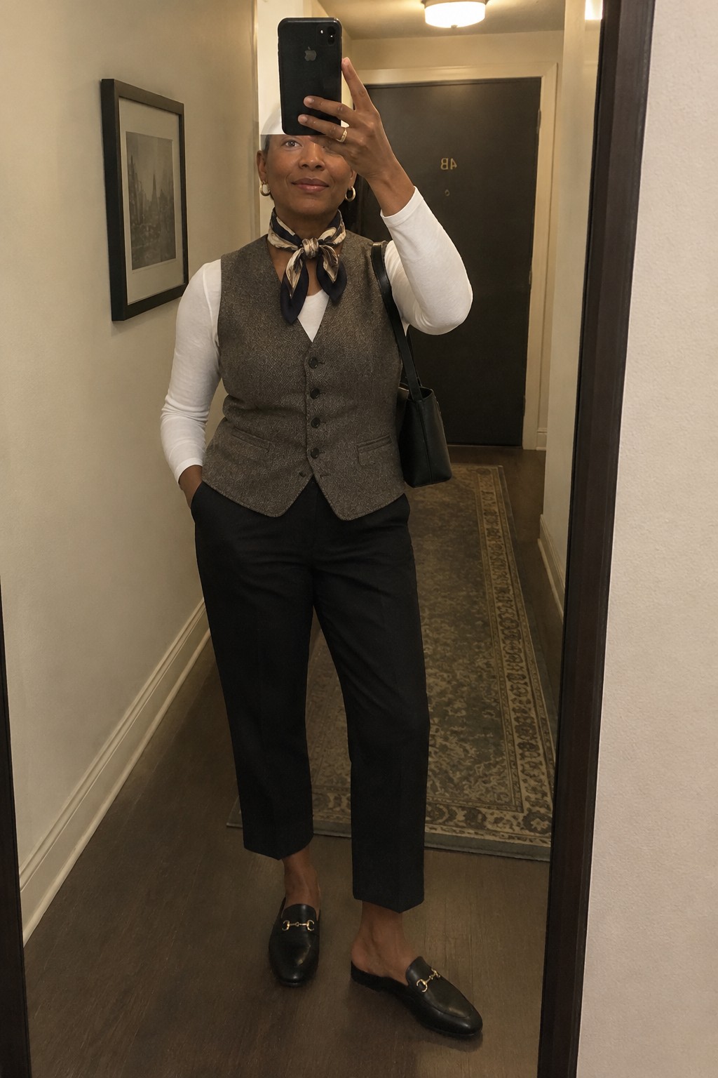 Woman in tweed vest, scarf, black pants, and loafers taking hallway mirror selfie