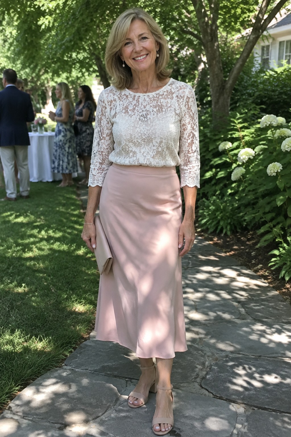 Smiling woman in white lace top and blush pink skirt holding clutch in garden