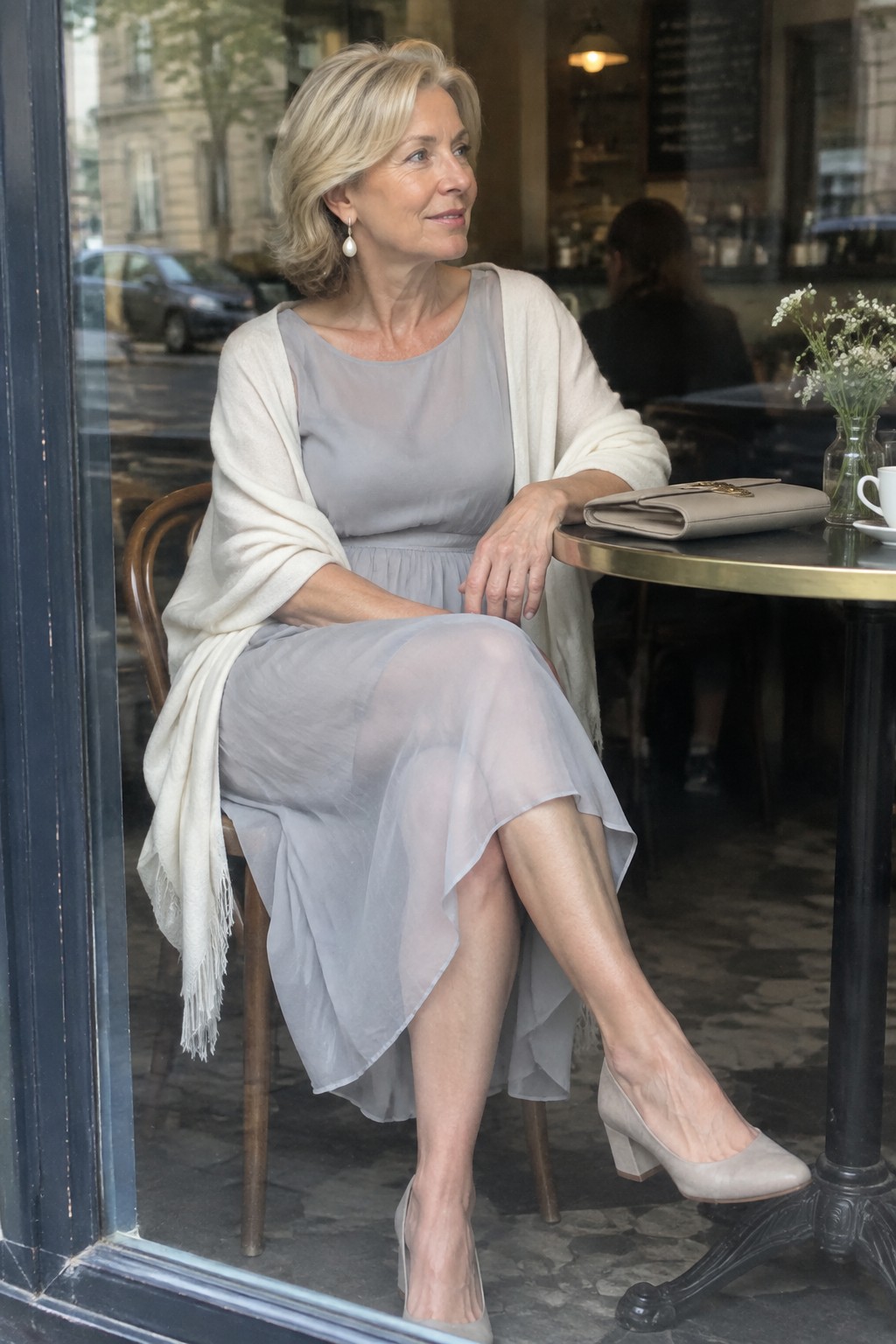 Blonde woman in sheer gray dress and white shawl seated at cafe table by window