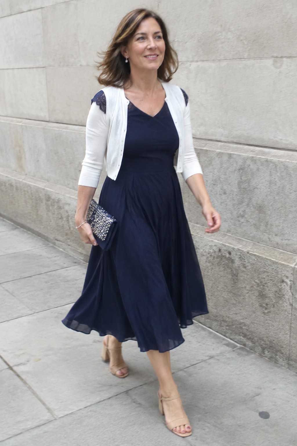 Mature woman in navy midi dress with white lace-trimmed cardigan, beige block heels, sparkly navy clutch, and delicate bracelet on stone pavement.
