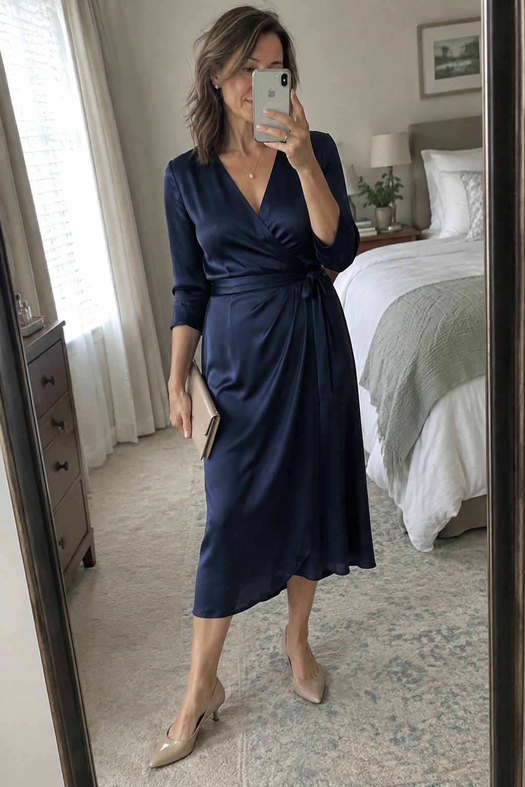 Woman in navy blue satin wrap dress taking bedroom mirror selfie with phone and clutch