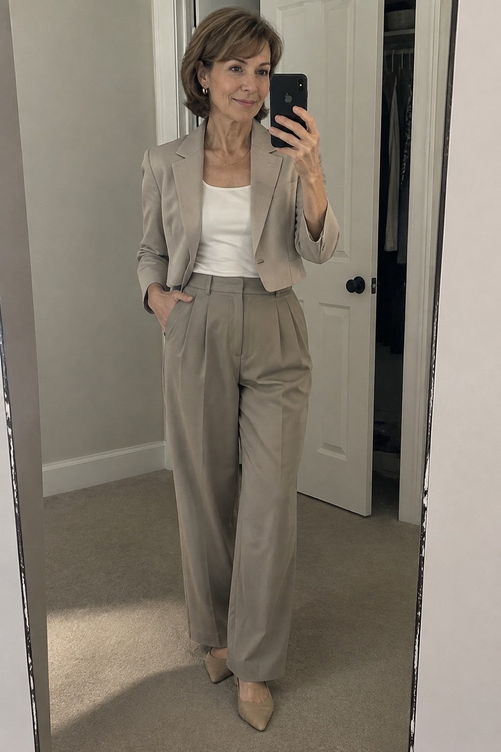 Woman in beige pantsuit holding phone for mirror selfie in hallway